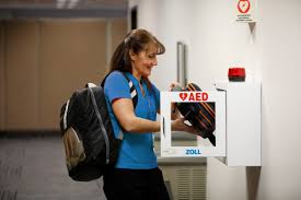 A woman is accessing the AED from it's wall cabinet and will perform an AED inspection.